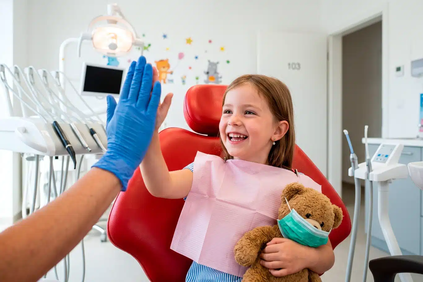 dentist child interaction Child in dentist chair giving high five holding teddy bear smiling